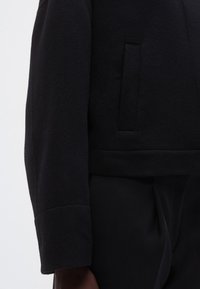 Veste en mélange de laine noire avec une finition texturée. Dotée d'un design court, de manches larges et d'une poche latérale cachée pour plus de fonctionnalité.