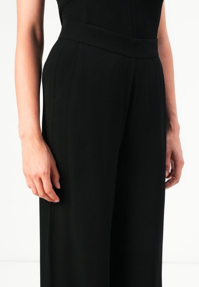 Person wearing high-waisted, wide-leg black trousers with hands relaxed at sides against a plain background.