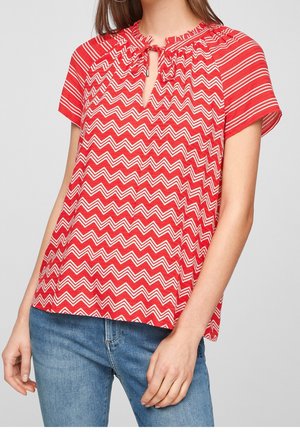 Kvinde iført en rød kortærmet top med hvide zigzag- og stribemønstre, nøglehulsudskæring med bindedetalje, kombineret med blå jeans.