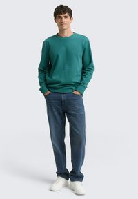 Felpa verde con maniche lunghe, vestibilità morbida, scollatura a girocollo. Abbinata a jeans in denim blu e sneakers bianche. Design semplice e casual.