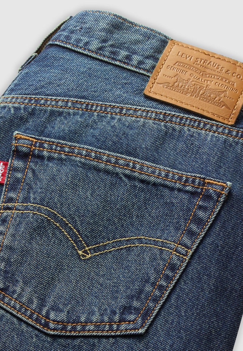 Poche en jean bleu avec des surpiqûres oranges contrastantes, une étiquette en cuir avec un logo en relief, et un design classique en V.