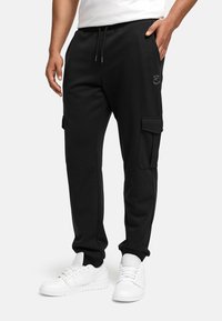 Pantalones cargo negros con cintura ajustable por cordón, bolsillos laterales y una textura suave. Cuenta con un logo sutil en la cadera izquierda. Lleva puestos unos zapatillas blancas.