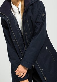 Dark blue parka jacket with a hood, featuring zip pockets, textured fabric, and silver hardware. Includes a contrasting inner lining.