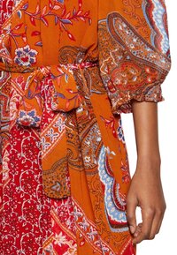 En kvinnas arm bredvid en tygbälte knutet i en rosett på en flerfärgad klänning med orange, röd, blå blommönster och paisley-mönster med puffärm.