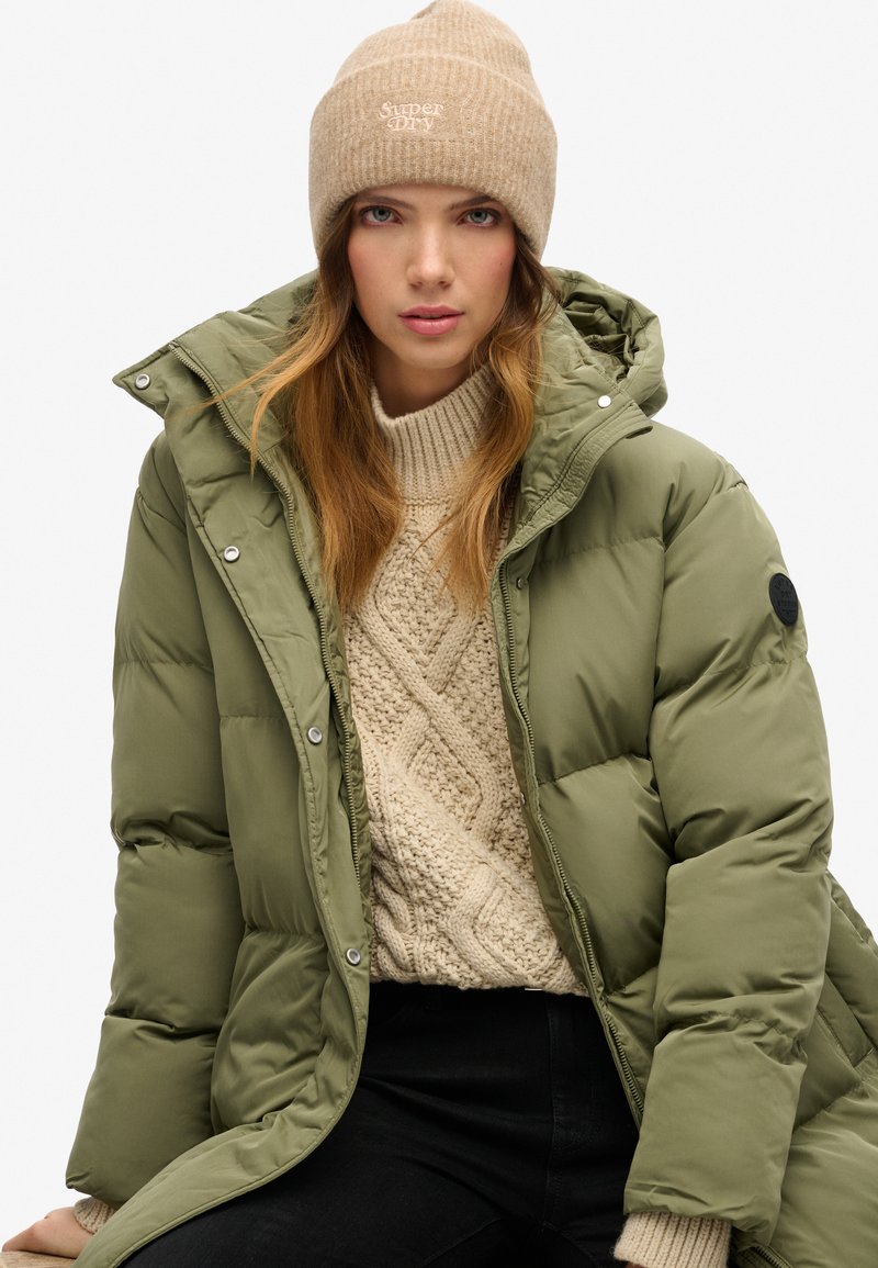 Olijfgroen gewatteerde jas met capuchon, voorzien van zilveren drukknoopsluitingen, gecombineerd met een crèmekleurige kabelgebreide trui en een beige geribbelde beanie met logo.