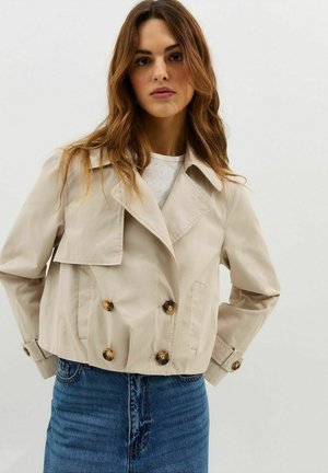 Vrouw met lang golvend haar, gekleed in een beige cropped double-breasted trenchcoat en een blauwe denim rok, staand tegen een effen witte achtergrond.