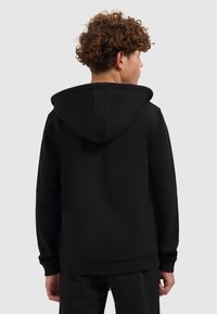 Jongere met krullend haar, gezien van achteren, in een zwarte hoodie en bijpassende zwarte broeken tegen een effen achtergrond.