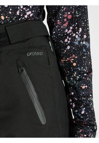 Zbliżenie na czarne spodnie marki Protest z zamkniętą kieszenią i regulowanym paskiem, w połączeniu z długim rękawem bluzką z wielokolorowym wzorem plam farby.