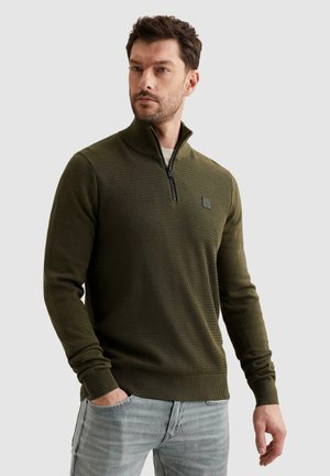 Man draagt een olijfgroen gestructureerd half-ritssluiting trui en lichtblauwe spijkerbroek, staand met één hand in de zak tegen een effen achtergrond.