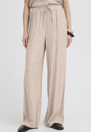 Béžové a biele široké nohavice s vertikálnymi pruhmi a šnúrkou v páse, nosené s dvoma náramkami na pravej zápästí a čiernymi špicatými topánkami.