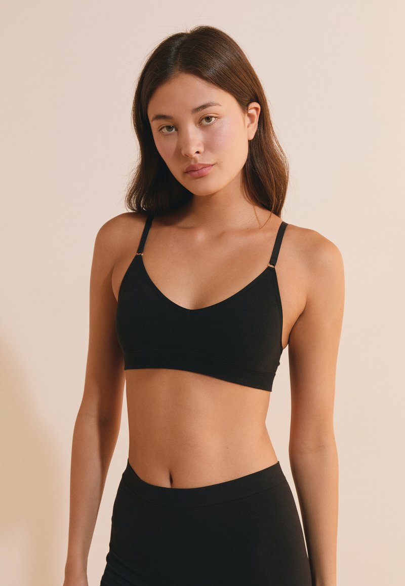 Giovane donna con lunghi capelli castani che indossa un bralette nero e slip neri a vita alta su uno sfondo beige.