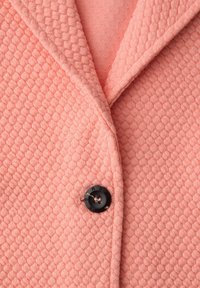 Gros plan sur une veste en tricot rose texturée avec un revers cranté et un seul bouton noir fermé à l'avant.