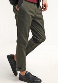 Homme portant un pantalon olive avec des poignets, des baskets noires, une main dans sa poche, se tenant contre un fond blanc.