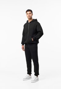 Zwarte hoodie met rits, voorzakken en geribbelde manchetten, gecombineerd met bijpassende zwarte joggers en witte sneakers op een effen achtergrond.