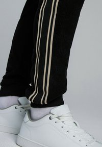 Czarne dresy z teksturowanej tkaniny i dwoma beżowymi paskami po bokach, w połączeniu z białymi sneakersami. Całość uzupełniają białe skarpetki.