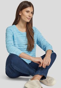 Hellblaues Langarmoberteil mit strukturiertem Wellenmuster, Rundhalsausschnitt, kombiniert mit dunklen Jeans und hellen Turnschuhen. Lässige Sitzpose.