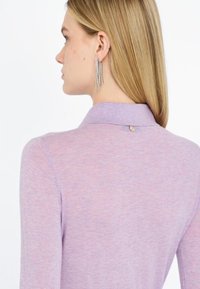 Polo shirt di un viola chiaro con colletto, caratterizzato da un bottone decorativo sul retro e un materiale morbido e testurizzato. Maniche lunghe visibili.