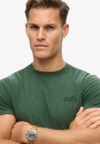 Groene katoenen t-shirt met korte mouwen en een ronde halslijn; heeft een klein geborduurd logo op de borst. Model draagt een tan kleurige horloge.