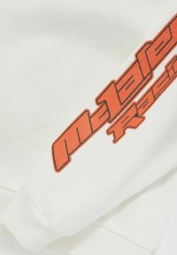 Bela majica s kapuco z oranžno teksturo "m1a1 racing" na rokavu. Ima rebraste manšete in mehak, gladek material.