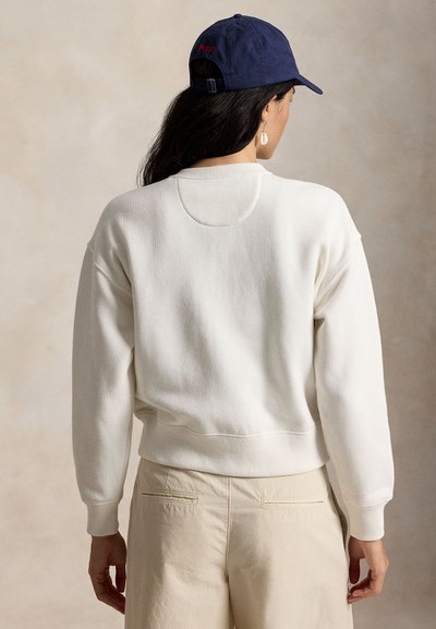 Γυναίκα που φοράει μπλε σκουφάκι, λευκό πουλόβερ με μακριά μανίκια και μπεζ παντελόνι, γυρισμένη με την πλάτη προς ουδέτερο φόντο.