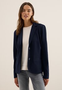 Kvinna iklädd en marinblå blazer över en krämfärgad skjorta och grå jeans, stående mot en enfärgad beige bakgrund.