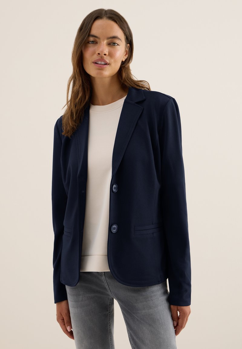Kvinna iklädd en marinblå blazer över en krämfärgad skjorta och grå jeans, stående mot en enfärgad beige bakgrund.