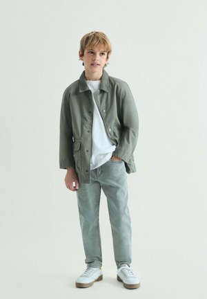 Niño con cabello rubio que lleva una chaqueta verde claro, camiseta blanca, pantalones verde claro y zapatillas blancas, de pie con una mano en el bolsillo.