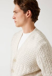 Helle beige Strickjacke mit strukturiertem Muster, zwei cremefarbenen Knöpfen und einem tiefen V-Ausschnitt. Weiches, gemütliches Material.