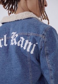 Jeans jakna s krznim ovratnikom, s bijelim natpisom "Kani" na leđima. Tekstura plave tkanine s vidljivim detaljima šivanja.