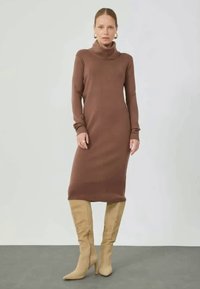 Brūna golfa, garām piedurknēm kleita no ribota auduma, ceļa garumā. Modelis valkā bēšas augstās zābaki līdz augšstilbam. Vienkārša silueta, bez rakstiem.