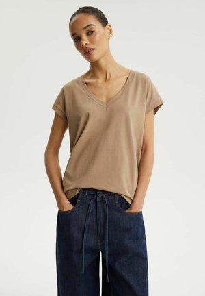 Kobieta ubrana w beżową bluzkę z krótkim rękawem i dekoltem w serek oraz luźne ciemnoniebieskie dżinsy, stojąca z rękami w kieszeniach na jasnym, gładkim tle.