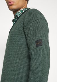 Pull vert foncé avec un tricot texturé. Comprend une écusson en cuir étiqueté « BOSS » sur la manche gauche. Chemise à col avec un intérieur à motif visible.
