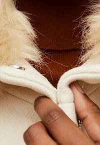 Chaqueta color crema con cuello de piel sintética, que presenta un cierre de cremallera y botón. Se puede ver una camiseta marrón de canalé debajo.