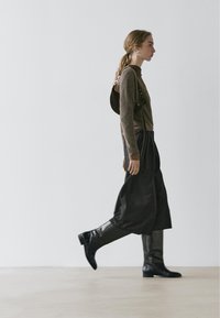Haut en maille marron, pantalon noir à jambes larges et bottes noires montantes jusqu'aux genoux. Cheveux longs coiffés en tresse, avec un sac à main simple porté à l'épaule et de grandes boucles d'oreilles.