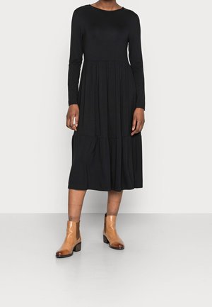 Kobieta w czarnej, długorękawowej sukience midi, w połączeniu z beżowymi botkami na tle jednolitego jasnoszarego tła.