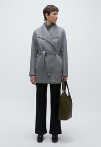 Γκρι παλτό τύπου wrap με ζώνη στη μέση, ριμπ γιακά και πλαϊνές τσέπες, συνδυασμένο με σκουρόχρωμο τζιν καμπάνα και τσάντα tote σε ελαιώδες πράσινο.