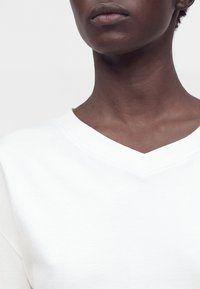 Gros plan sur le cou et les lèvres d’une personne portant un t-shirt blanc à col en V sur un fond clair uni.