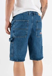 Pantalones cortos de denim en azul medio, con múltiples bolsillos grandes, detalles de costura y dobladillos deshilachados, con un acabado texturizado natural.