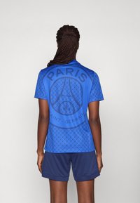 Blå sportströja med geometriskt mönster, med "PARIS" tydligt på ryggen, parat med marinblå shorts. Slät textur, modern design.