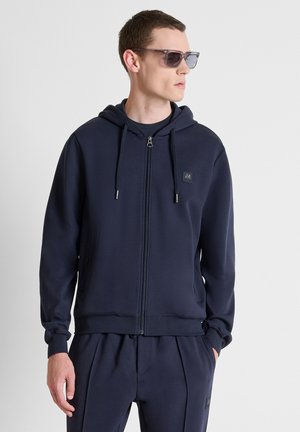 Man draagt een donkerblauwe hoodie met rits en bijpassende joggingbroek, met een zonnebril, kijkt naar rechts tegen een effen achtergrond.