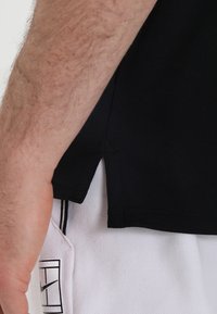 Camiseta atlética negra de manga corta, con un dobladillo lateral. Llevada sobre pantalones cortos blancos con franjas de acento negras y logotipo.