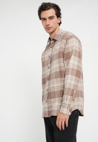 Brun rutete skjorte med hvite linjer; har lange ermer og knapper på mansjettene; laget av vevd stoff og har spiss krage.