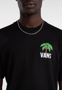 Camiseta negra de manga corta con un gráfico de palmera verde y el texto "VANS" en el lado izquierdo del pecho. Accesorios con un collar de cadena plateada.