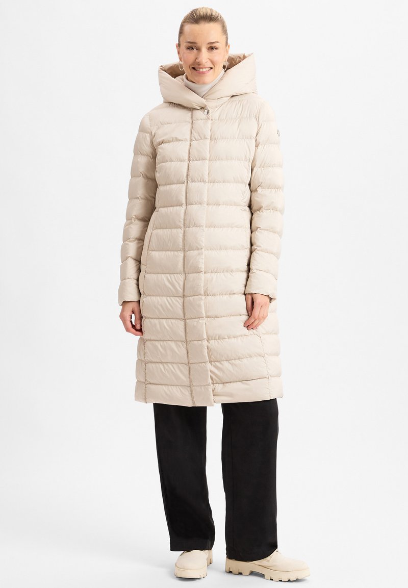Beige Daunenjacke mit Kapuze, mit horizontalem Steppdesign, mittigem Reißverschluss und einer lockeren Passform. Getragen über schwarzen Hosen und cremefarbenen Schuhen.