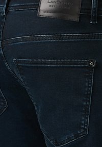 Ciemnoniebieskie dżinsy mają tylną kieszeń z widocznym przeszyciem oraz skórzaną naszywkę z logo na pasku. Teksturowana tkanina o gładkim wykończeniu.