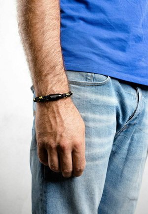 Pulsera trenzada negra y amarilla en una muñeca, con un diseño texturizado y un acento central sólido, llevada con una camisa azul y jeans.