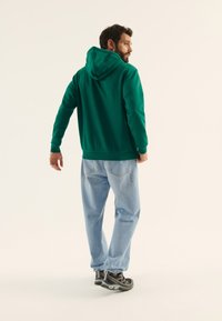 Sudadera verde con bolsillo tipo canguro, puños y dobladillo acanalados, combinada con jeans de corte holgado en azul claro y zapatillas gris. Estilo y textura minimalista.