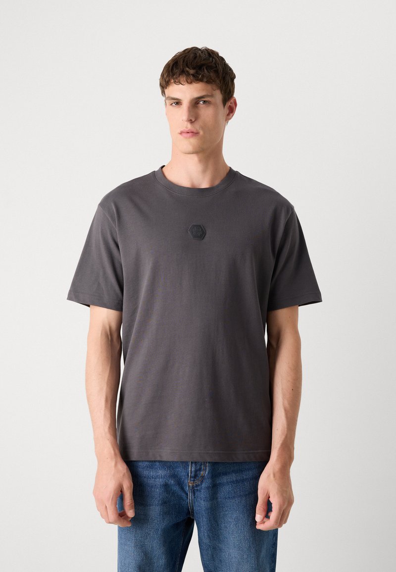 T-shirt en coton gris à manches courtes, col rond et logo hexagonal noir au centre. Porté avec un jean en denim bleu.