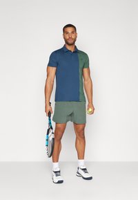 Tumesinine ja roheline lühikeste varrukatega polo särk, millele on sobitatud rohelised lühikesed püksid. Model hoiab tennisereketit ja palli, jalas valged spordijalatsid.