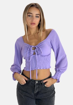 Lavendelfarbene, kürzere Bluse mit gerafftem Bund, Bindedetail am Ausschnitt und langen Puffärmeln, kombiniert mit hochgeschnittenen schwarzen Jeans.